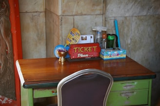 A wooden table with a dark green base, featuring a variety of objects, is set against a marble wall. On the table is a red sign with yellow lettering that reads 'TICKET Please.' Next to the sign, there's a small brass bowl containing folded papers, a colorful tissue box with a beach motif, and a plastic bottle. In the background, a fan, cleaning supplies, and a flyer are visible.