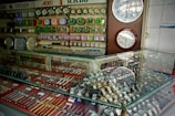 A display case filled with various types of wristwatches, organized with metal bands and different colors. Wall clocks are mounted on the wall behind the case, along with a range of colorful alarm clocks arranged in rows. Brand names like SEIKO, RADO, and CITIZEN are visible, suggesting a retail setting for timepieces.