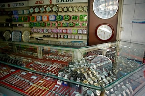 A display case filled with various types of wristwatches, organized with metal bands and different colors. Wall clocks are mounted on the wall behind the case, along with a range of colorful alarm clocks arranged in rows. Brand names like SEIKO, RADO, and CITIZEN are visible, suggesting a retail setting for timepieces.