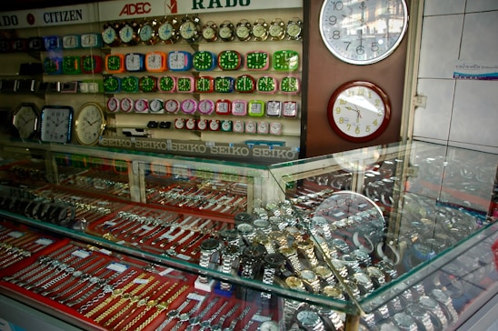 A display case filled with various types of wristwatches, organized with metal bands and different colors. Wall clocks are mounted on the wall behind the case, along with a range of colorful alarm clocks arranged in rows. Brand names like SEIKO, RADO, and CITIZEN are visible, suggesting a retail setting for timepieces.