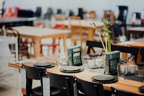 A well-arranged dining table set in what appears to be a showroom or a store, featuring black chairs and a wooden table. The table is adorned with black plates, transparent glasses, white cups, and two vases with tall, spiraled green plants. Price tags hang from the chairs, and other tables and chairs are visible in the blurred background.