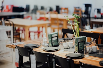 A well-arranged dining table set in what appears to be a showroom or a store, featuring black chairs and a wooden table. The table is adorned with black plates, transparent glasses, white cups, and two vases with tall, spiraled green plants. Price tags hang from the chairs, and other tables and chairs are visible in the blurred background.