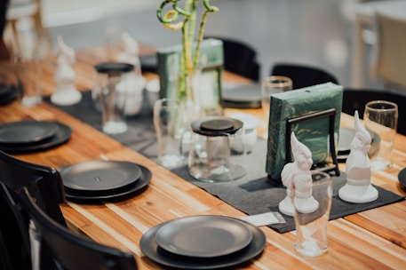 A dining table is elegantly set with black plates, clear glass cups, and decorative objects, including white figurines. The table runner is dark, and there are green book-like items standing upright at the center. The warm-toned, wooden surface adds an earthy touch to the setup.