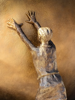 A bronze sculpture depicts a person from behind, reaching up with both hands against a textured golden surface. The detailed artistry emphasizes the figure's outstretched arms and the textured pattern of their clothing.