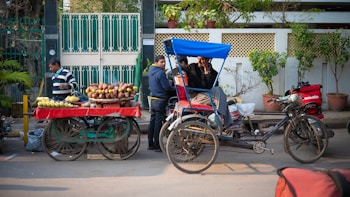 A street scene featuring a vendor with a cart laden with corn and bananas and a cycle rickshaw with people seated inside. A delivery person with a red bag is seen on a scooter, and another man is wearing a striped sweater. The background includes a green and white fence and some potted plants.