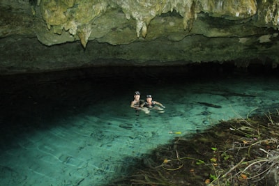 Two people wearing swim goggles and snorkels are floating in clear turquoise water inside a cave. The cave features rock formations with stalactites hanging from the ceiling. The surroundings are calm, and the water is transparent, reflecting the rocky roof above.
