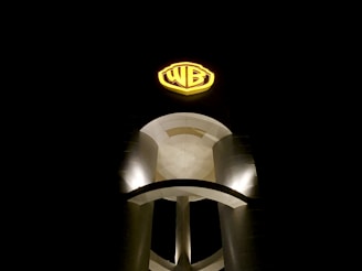 A large, illuminated sign featuring the letters 'WB' is prominently displayed against a dark night sky. The sign is positioned on a modern, cylindrical structure with smooth curves and is highlighted by bright lighting.