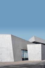 A modern building features minimalist architecture with large concrete panels and sharp angles, set against a clear blue sky. The structure includes reflective windows and angular shapes that add to its contemporary design.