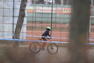 A child wearing a helmet rides a bright orange mountain bike on a path next to a chain-link fence. The fence has a sports field in the background with red dirt. The area is bounded by a blue ribbon, likely indicating a designated biking path.
