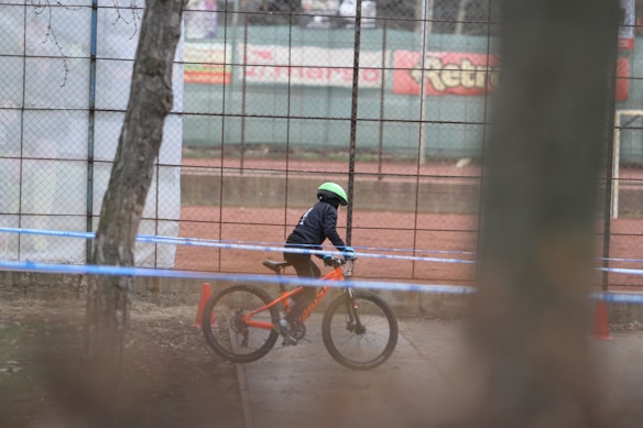 A child wearing a helmet rides a bright orange mountain bike on a path next to a chain-link fence. The fence has a sports field in the background with red dirt. The area is bounded by a blue ribbon, likely indicating a designated biking path.