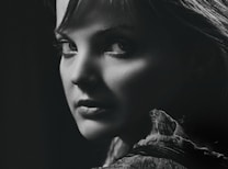 A black and white portrait of a person with a captivating expression, featuring dramatic lighting that highlights the contours of their face and the texture of their clothing. The focus is on the eyes, creating a mysterious and intense mood.