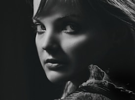 A black and white portrait of a person with a captivating expression, featuring dramatic lighting that highlights the contours of their face and the texture of their clothing. The focus is on the eyes, creating a mysterious and intense mood.