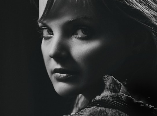 A black and white portrait of a person with a captivating expression, featuring dramatic lighting that highlights the contours of their face and the texture of their clothing. The focus is on the eyes, creating a mysterious and intense mood.