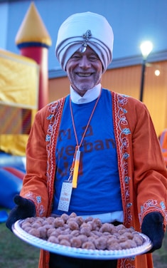 A smiling person wearing a white turban and an orange embroidered coat holds a tray of small brown pastries. The individual is also wearing a blue T-shirt and a name tag labeled 'Fall Festival.' In the background, there is an inflatable play structure and a softly lit environment typical of a festival setting.