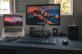 A home office setup with a laptop and a desktop monitor displaying the same mountainous landscape wallpaper. A keyboard and mouse are placed on the black desk, alongside two small speakers and a smartphone. Natural light filters through the window in the background.