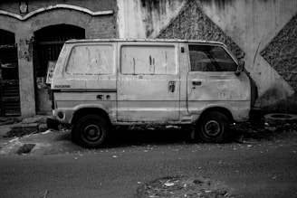An old, worn-out van with visible dirt and damage is parked next to a textured, dilapidated wall. The scene appears to be in an urban environment, with debris and litter scattered on the ground.