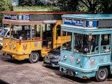 Two colorful tourist buses are parked side by side with visible branding for the Bandung city tour, one in a bright yellow color and the other in turquoise. A black car is partially visible between the buses. The scene is set outdoors with trees providing shade in the background.