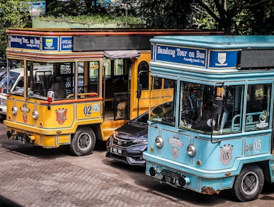 Two colorful tourist buses are parked side by side with visible branding for the Bandung city tour, one in a bright yellow color and the other in turquoise. A black car is partially visible between the buses. The scene is set outdoors with trees providing shade in the background.