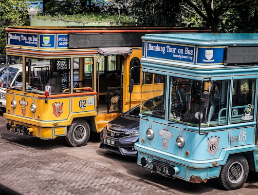 Two colorful tourist buses are parked side by side with visible branding for the Bandung city tour, one in a bright yellow color and the other in turquoise. A black car is partially visible between the buses. The scene is set outdoors with trees providing shade in the background.