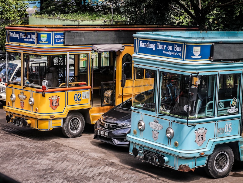 Two colorful tourist buses are parked side by side with visible branding for the Bandung city tour, one in a bright yellow color and the other in turquoise. A black car is partially visible between the buses. The scene is set outdoors with trees providing shade in the background.