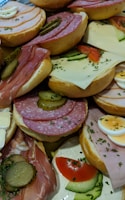 A variety of open-faced sandwiches featuring an assortment of meats, cheeses, and toppings. Slices of salami, ham, and prosciutto are paired with cheese such as Swiss and Brie, adorned with fresh vegetables like cucumbers, tomatoes, and sliced boiled eggs. Pickles and a sprinkling of herbs garnish the sandwiches, which are arranged closely together on a platter.