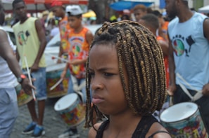 A group of people are playing percussion instruments in an outdoor setting. The focus is on a young person with braided hair looking serious or contemplative. Surrounding them are others engaged in drumming, wearing colorful clothing with printed designs. The background shows various blurred figures and elements indicating a lively street or festival environment.