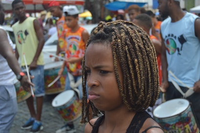 A group of people are playing percussion instruments in an outdoor setting. The focus is on a young person with braided hair looking serious or contemplative. Surrounding them are others engaged in drumming, wearing colorful clothing with printed designs. The background shows various blurred figures and elements indicating a lively street or festival environment.