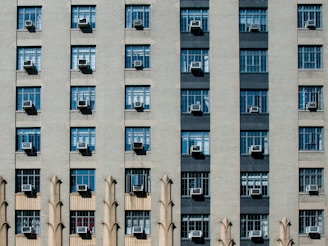 A symmetrical building facade with multiple rows and columns of windows, each featuring air conditioning units. The architecture incorporates decorative elements between some of the windows, adding a repetitive geometric pattern.