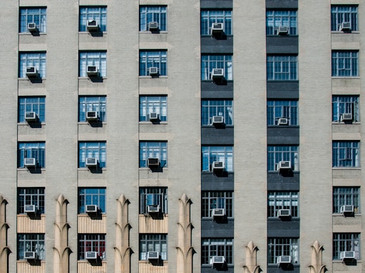 A symmetrical building facade with multiple rows and columns of windows, each featuring air conditioning units. The architecture incorporates decorative elements between some of the windows, adding a repetitive geometric pattern.
