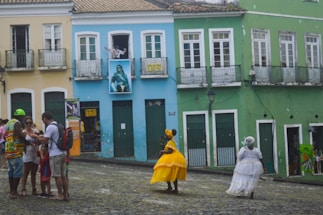 A cobblestone street in front of colorful Colonial-style buildings with pastel yellow, blue, and green facades. Several people are gathered, some wearing traditional, colorful dresses and others in casual clothing. One person is waving from a window above, and the street has a lively, cultural atmosphere.