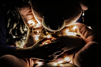 A large teddy bear is illuminated by a string of warm fairy lights wrapped around it. A child's hands are seen gently holding the teddy bear's paw, creating a cozy and intimate atmosphere.