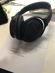 On a desk lies a pair of black over-ear headphones resting on a paper with mathematical text. Nearby, there are open books, part of a smartphone, and writing instruments, suggesting a study or work environment.