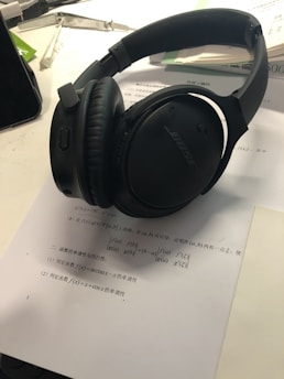 On a desk lies a pair of black over-ear headphones resting on a paper with mathematical text. Nearby, there are open books, part of a smartphone, and writing instruments, suggesting a study or work environment.