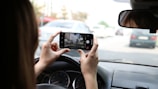 Person holding a smartphone while sitting in the driver's seat of a car, taking a photo or video of traffic ahead.