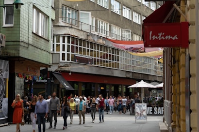 A busy street scene with numerous people walking along a pedestrian area lined with shops. The buildings display a mix of architectural styles, with modern storefronts and older, worn facades. Signs and awnings in various colors hang over the shops, indicating a commercial area. People appear to be casually dressed, suggesting a relaxed urban environment.