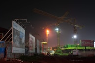 A nighttime construction site illuminated by artificial lights. Two large cranes tower over the partially constructed building. Billboards flank the left side of the image, displaying advertisements. The background features a misty sky with dim lighting accentuating the urban setting.