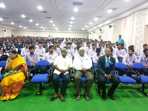 A large auditorium is filled with seated students dressed in uniforms, most of whom are wearing lanyards with identification cards. At the front, there are several individuals, including an older woman in a yellow saree and three men, sitting in blue chairs. The room is well-lit with ceiling lights and has decorated walls.