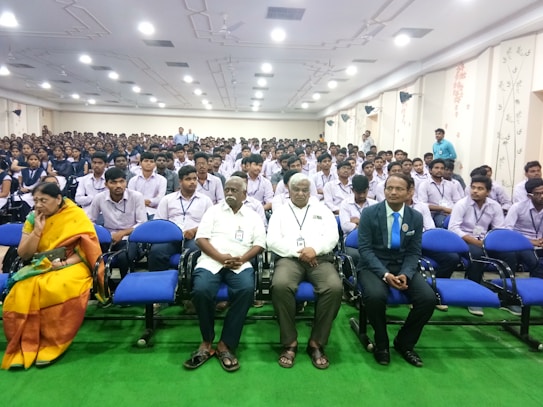 A large auditorium is filled with seated students dressed in uniforms, most of whom are wearing lanyards with identification cards. At the front, there are several individuals, including an older woman in a yellow saree and three men, sitting in blue chairs. The room is well-lit with ceiling lights and has decorated walls.