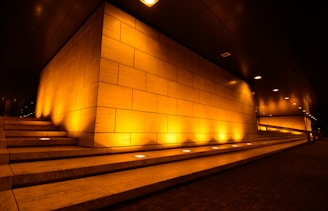A corner of a modern building is highlighted by ambient yellow-orange lighting. The walls are composed of large rectangular tiles, and there are three sets of steps leading up to the structure. The lighting creates an inviting and warm atmosphere, contrasting with the dark ceiling above.