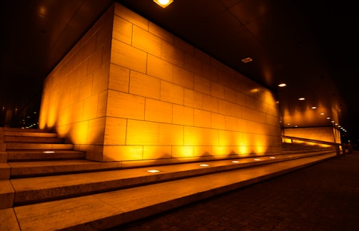 A corner of a modern building is highlighted by ambient yellow-orange lighting. The walls are composed of large rectangular tiles, and there are three sets of steps leading up to the structure. The lighting creates an inviting and warm atmosphere, contrasting with the dark ceiling above.