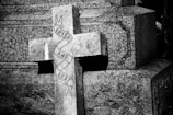 A weathered stone cross with the words 'Loved and Lost' wrapped around it, set against a textured stone background. The image is cast in black and white, highlighting the somber tone.
