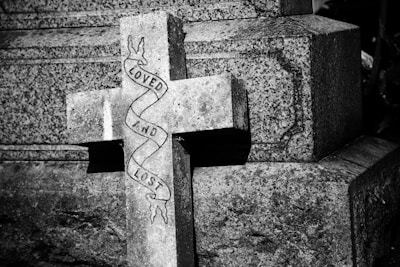 A weathered stone cross with the words 'Loved and Lost' wrapped around it, set against a textured stone background. The image is cast in black and white, highlighting the somber tone.