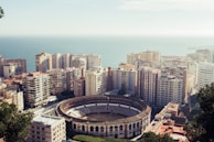 An aerial view of a coastal city featuring a large, circular bullring surrounded by tall apartment buildings. The cityscape extends towards the ocean, which is visible in the background. The buildings are densely packed, displaying various colors and architectural styles.