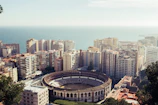An aerial view of a coastal city featuring a large, circular bullring surrounded by tall apartment buildings. The cityscape extends towards the ocean, which is visible in the background. The buildings are densely packed, displaying various colors and architectural styles.