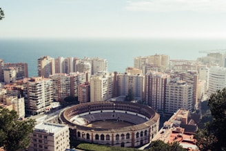 An aerial view of a coastal city featuring a large, circular bullring surrounded by tall apartment buildings. The cityscape extends towards the ocean, which is visible in the background. The buildings are densely packed, displaying various colors and architectural styles.