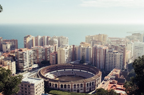 An aerial view of a coastal city featuring a large, circular bullring surrounded by tall apartment buildings. The cityscape extends towards the ocean, which is visible in the background. The buildings are densely packed, displaying various colors and architectural styles.