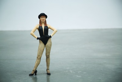 A standing woman dressed in a black leotard, fishnet stockings, and black gloves. She is wearing a black hat and posed confidently with her hands on her hips on a gray concrete floor with a plain white wall as a backdrop.