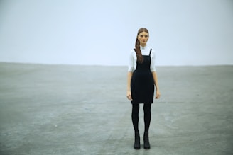 A woman with long hair stands in a minimalist setting, wearing a black pinafore dress over a white blouse. She is positioned in the center of the image with her arms relaxed by her sides. The floor is a smooth, gray concrete, and the background is a plain, light gray wall.