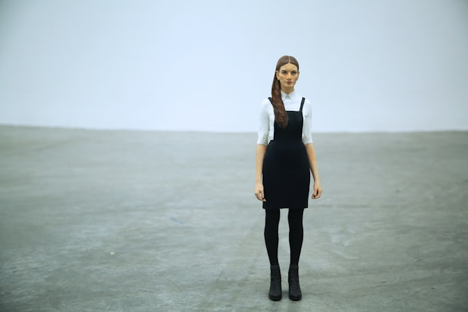 A woman with long hair stands in a minimalist setting, wearing a black pinafore dress over a white blouse. She is positioned in the center of the image with her arms relaxed by her sides. The floor is a smooth, gray concrete, and the background is a plain, light gray wall.