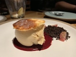 A dessert plate features a creamy mousse-like dessert topped with a thin, crispy caramelized layer. The dessert sits atop a pool of dark red sauce, accompanied by a small pile of crumbled dark chocolate and some sliced fruit garnishes, possibly grapes. The setting suggests a refined dining atmosphere.